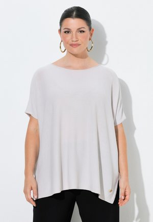 Kvinde med mørkt hår iført en løs hvid kortærmet top og sorte bukser, bærende store gyldne hoops-øreringe, stående mod en hvid baggrund.