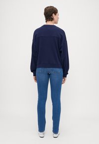 Sudadera azul marino con cuello redondo y hombros caídos, combinada con vaqueros ajustados azules y zapatos plateados, vista desde atrás.