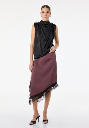 Femme portant un haut en satin noir sans manches et une jupe asymétrique mauve avec une bordure en dentelle noire, assortie à des talons ouverts noirs.