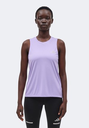 Kvinde stående med fronten mod kameraet iført en ærmeløs lavendel atletisk top og sorte leggings med hvide striber på en ensartet lys baggrund.