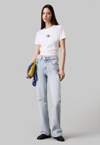 Camiseta blanca corta con logo, vaqueros acampanados azul claro, sosteniendo un bolso amarillo y un suéter morado. Zapatos negros de punta. Diseño sencillo.