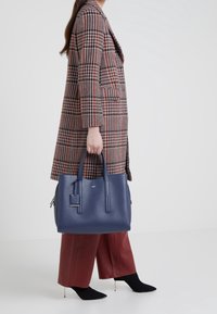 Osoba ubrana w płaszcz w jodełkę i szerokie czerwone spodnie, trzymająca dużą granatową skórzaną torbę, stojąca na białej podłodze.