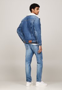 Giacca di denim con colletto in pelliccia sintetica bianca, tessuto blu, chiusura con bottoni e patch in pelle con marchio sul retro. Indossata con jeans azzurri.