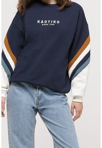 Felpa navy con strisce bianche, arancioni e teal sulle maniche; presenta il logo "KAOTIKO" e una vestibilità rilassata. Abbinata a jeans azzurri.