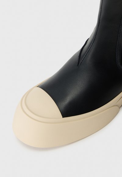 Černá kožená bota s béžovou gumovou podešví a kulatou špičkou, s prošíváním a elastickými bočními panely, na bílém pozadí.