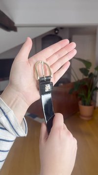 Juodas odinis diržas, laikomas rankoje, su auksiniu metaliniu dvigubu dantyto sagties spaustuku ir dekoratyviniu kilpeliu šalia sagties. Lygus paviršius.