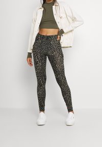 Hochtaillierte Leggings mit einem dunkelgrünen und schwarzen Leopardenmuster. Weicher, dehnbarer Stoff mit glatter Textur, kombiniert mit einem enganliegenden grünen Oberteil und weißen Sneakers.