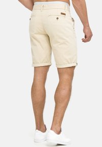 Beige katoenen shorts met opgerolde zomen, voorzien van achterzakken en knoopdetails, gestyled voor een casual pasvorm. Gecombineerd met witte sneakers.