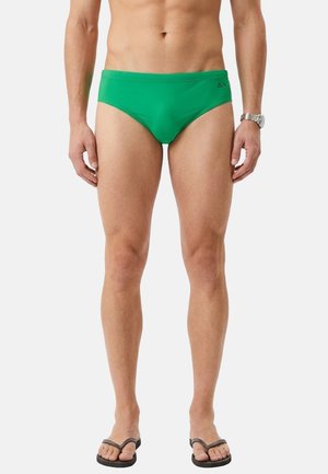 Uomo che indossa slip da bagno verdi, infradito nere e un orologio da polso argentato, in piedi contro uno sfondo chiaro e uniforme.