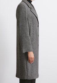 Abrigo largo de houndstooth en blanco y negro con cuello descuidad, dos bolsillos delanteros y una silueta recta, mostrado de lado.