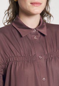 Camisa castanha de botão, com uma textura suave e macia, apresentando uma gola, detalhes franzidos e botões combinando na parte da frente.