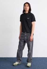 Camiseta negra con mangas enrolladas y logo verde; vaqueros grises con un patrón de ajuste suelto; zapatillas verdes y grises. Fondo liso.