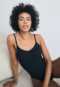 Ženska s kodrastimi lasmi, oblečena v črno brezrokavnico, sedi na svetlem teksturiranem preprogi, opira se na eno roko in gleda naravnost.