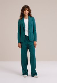 Súprava v tyrkysovej farbe obsahuje priliehavé sako s vykrojenými klopami a nohavice s širokými nohavicami. Nosená cez bielu košeľu, doplnená bielymi teniskami.