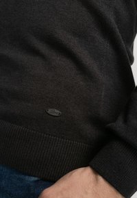 Donker gebreide trui met geribbelde zoom en manchet. Beschikt over een klein zwart logo-label nabij de zoom. Materiaal lijkt zacht en textuurrijk.