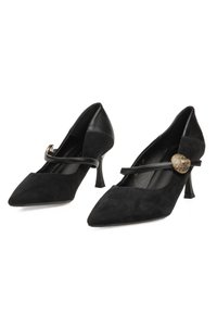 Tacones Mary Jane de ante negro con punta puntiaguda, tiras delgadas y hebillas con forma de corazón texturizadas en dorado, con detalles de cuero y tacones de aguja.
