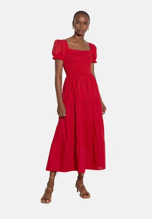 Kobieta w czerwonej, wielowarstwowej sukience midi z bufkami i kwadratowym dekoltem, w beżowych sandałach na obcasie z wiązaniami, stojąca na białym tle.