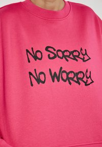 Felpa rosa con scollo rotondo, caratterizzata da testo in stile graffiti nero "No Sorry No Worry". Materiale morbido e vestibilità ampia.
