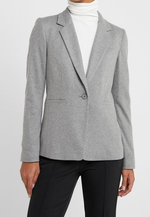 Femme portant un blazer gris clair à un bouton, un col roulé blanc et un pantalon noir ajusté sur un fond uni.
