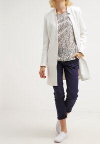 Blazer bianco lungo su una camicia con motivo, pantaloni blu navy e scarpe slip-on bianche. Il blazer presenta un design elegante con bottoni minimalisti.