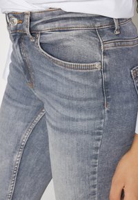 Vaqueros de denim azul de lavado medio con un ajuste ceñido, que presentan herrajes plateados, bolsillos delanteros y una sutil textura desgastada.