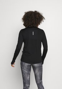 Black long-sleeve top with a high collar and thumbholes, featuring a thin vertical white stripe on the back, paired with patterned gray leggings.