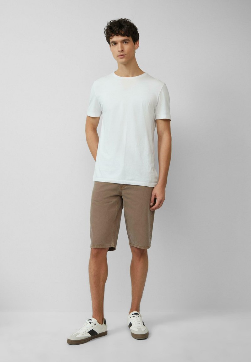 Camiseta blanca de algodón combinada con pantalones cortos de mezclilla marrón claro. Modelo lleva zapatillas blancas con detalles en negro. Diseño simple y casual.