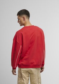 Felpa rossa con vestibilità ampia, scollo rotondo e spalle cadenti. Presenta una striscia laterale bianca e una texture di tessuto morbido.