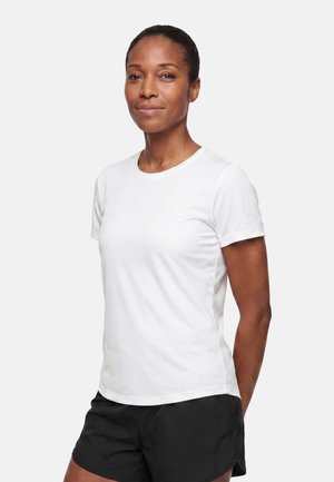 Femme aux cheveux courts portant un t-shirt blanc uni à manches courtes et un short noir, debout devant un fond blanc.