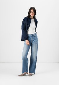 Marineblå jakke over en hvit skjorte, kombinert med lys blå brede jeans med frayed kanter og grå spisse sko.