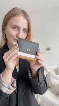 Melna teksturēta ādas maciņš ar zelta logo, apakšā ir balta un brūna paneļa kombinācija. Kompakts dizains ar magnētisku aizdari.