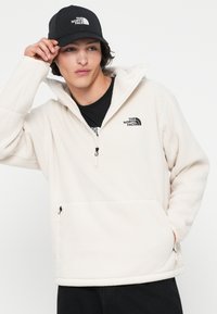 Giovane che regola un cappellino nero della North Face, indossando un fleece bianco con zip e tasca frontale e una maglietta nera su uno sfondo semplice.
