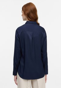 Marineblå satin skjorte med krave, lange ærmer og knappede manchetter. Har en central plissé og en rundet kant.