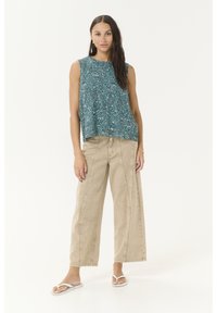 Frau mit langen dunklen Haaren, die ein ärmelloses, gemustertes türkisfarbenes Oberteil, weite beige Hosen und weiße Flip-Flops trägt, steht vor einem schlichten Hintergrund.