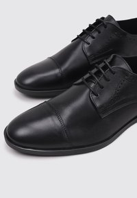 Zapatos de cuero negro con una textura suave, diseño de puntera cerrada y detalles de brogue. Cuentan con cordones y una silueta elegante y minimalista.