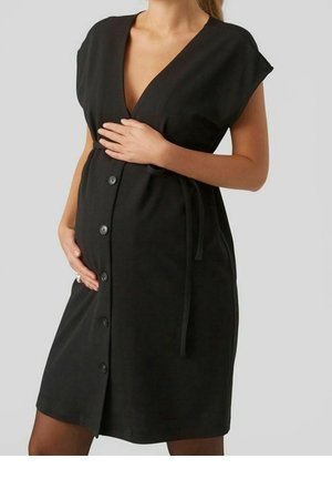 Osoba w ciąży w czarnej sukience zapinanej na guziki z dekoltem w serek i krótkimi rękawami, ręce delikatnie obejmujące brzuch na neutralnym tle.