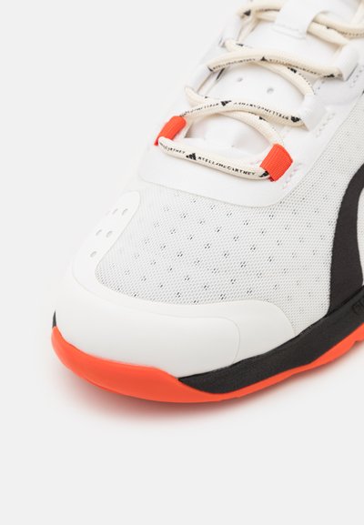 White athletic shoe with a textured mesh upper, orange accents, and a black sole. Features perforated detailing and light-colored laces.