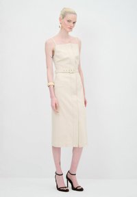 Robe midi couleur crème avec fines bretelles, corsage ajusté et taille ceinturée. Présente une coupe droite et des détails de couture avant subtils.