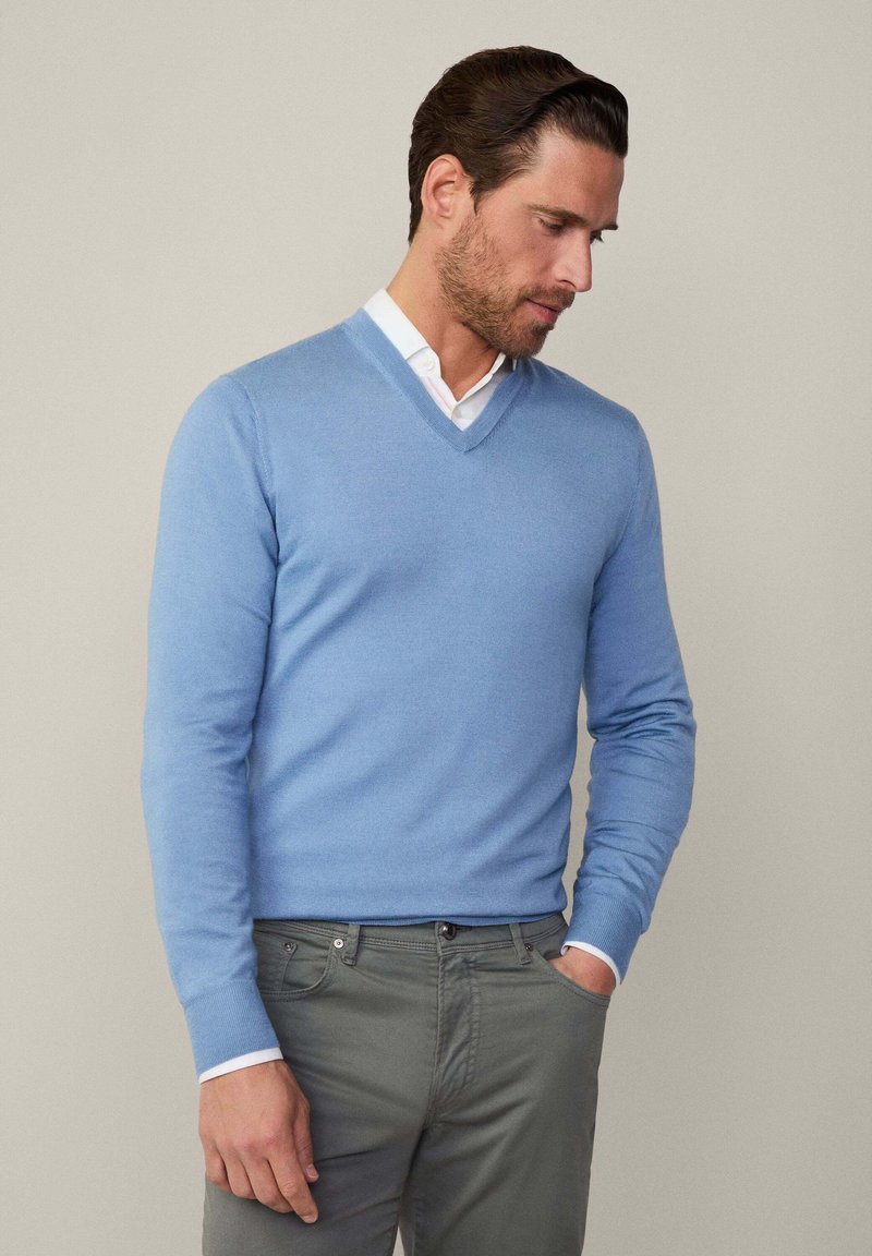 Hellblauer V-Ausschnitt-Pullover aus Strickstoff, mit langen Ärmeln und taillierter Passform, getragen über einem weißen Hemd mit Kragen.