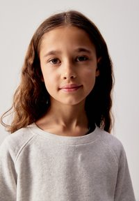 Jeune fille aux longs cheveux bruns portant un sweat-shirt gris clair, debout devant un fond clair uni, regardant vers la caméra.