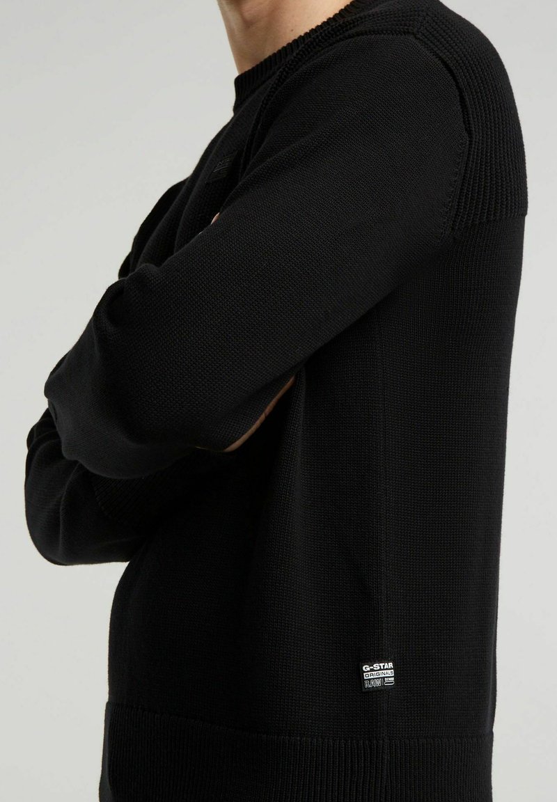 Suéter negro de punto de manga larga, usado con los brazos cruzados, con una pequeña etiqueta de G-Star Originals Raw cerca del dobladillo sobre un fondo liso.