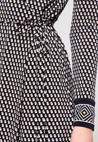 Black dress with white and beige polka dots, featuring a tied waist, long sleeves, and geometric patterned cuffs for added detail.