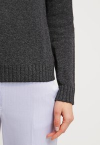 Ciemnoszary sweter z dzianiny z prążkowanymi mankietami i dołem, o miękkiej fakturze. Pod spodem widoczna jest jasna, w kratkę tkanina koszuli.