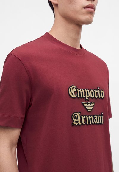 Млад мъж, носещ червена тениска с къс ръкав с бежов текст "Emporio Armani" и лого на гърдите.