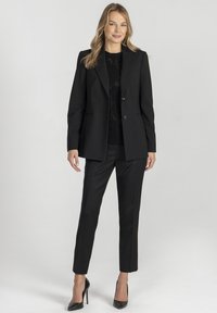 Czarny, dopasowany żakiet z pasującymi spodniami w fasonie slim fit, o gładkiej fakturze i z pojedynczym zapięciem na guzik, zestawiony z czarnymi szpilkami.