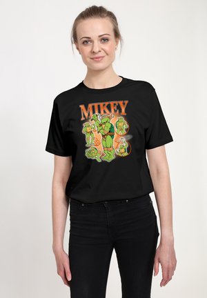 Kobieta nosząca czarną koszulkę z wieloma zielonymi postaciami Mikey z Żółwi Ninja oraz pomarańczowym napisem "MIKEY" u góry.
