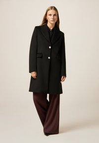 Cappotto di lana nera con vestibilità sartoriale, rever a punta e chiusure con bottoni dorati, abbinato a pantaloni ampi color bordeaux. Texture liscia e strutturata.