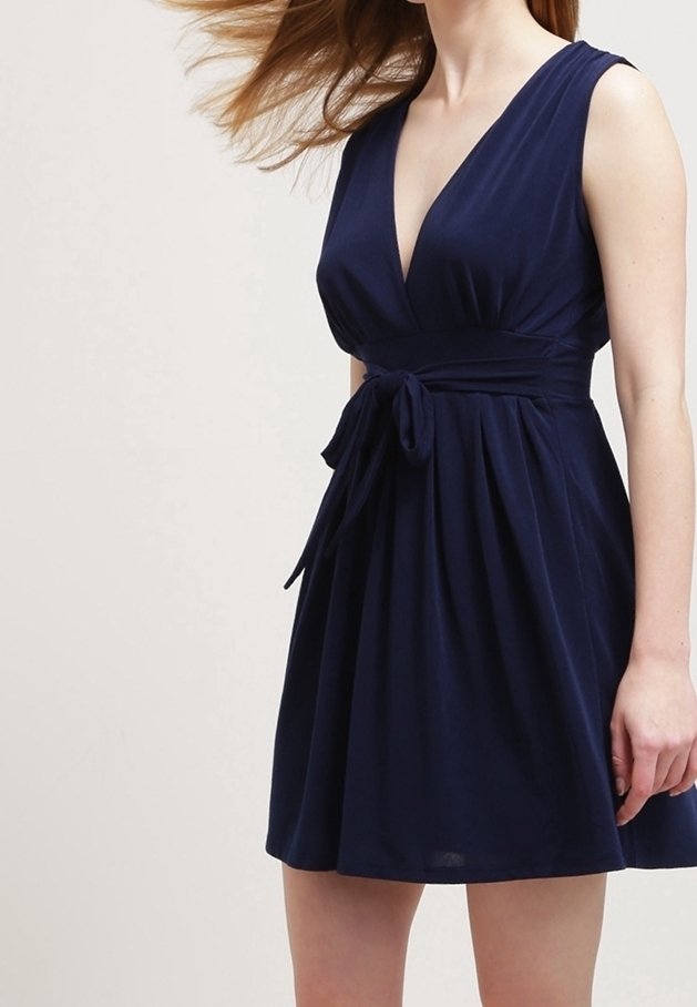 Femme portant une robe sans manches bleu marine avec un décolleté en V profond et une ceinture à la taille, les cheveux soufflés sur le côté sur un fond clair uni.