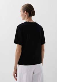 Zwarte katoenen t-shirt met korte mouwen en een ronde hals, met een ontspannen pasvorm. Van achteren bekeken, in combinatie met witte broeken.