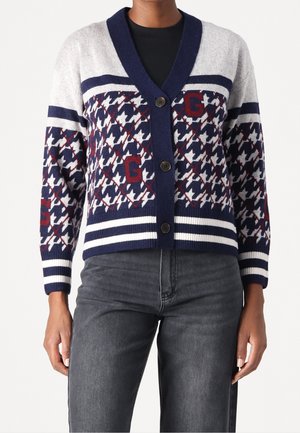 Personne portant un cardigan boutonné bleu marine, blanc et gris avec des motifs pied-de-poule et lettre G, associé à un jean gris foncé.